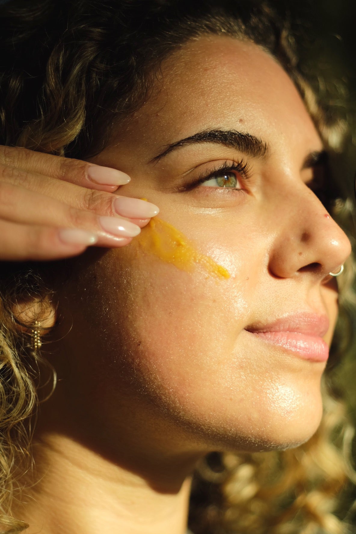 Close-up of a woman's face with a hand applying a yellow cream to her cheek.