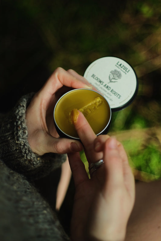 Person holding a small container of golden salve with Lazuli Remedies branding, outdoors.