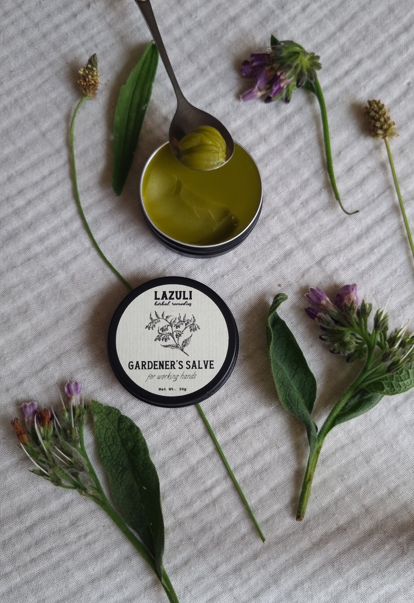Lazuli Gardener's Salve jar with a spoonful of salve and herbs on a textured surface