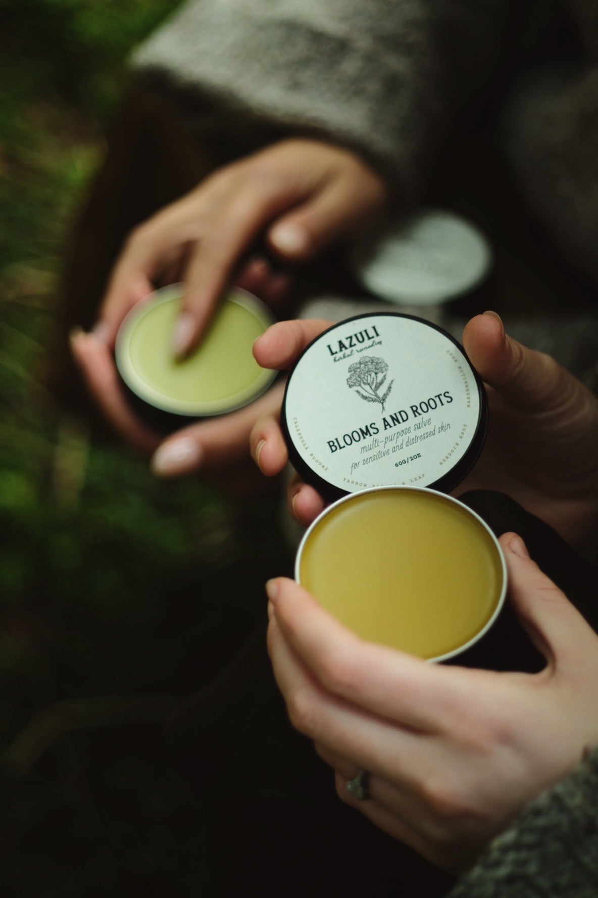 Two hands holding open tins of  Lazuli Blooms and Roots Muti-Purpose Healing Salve and Lazuli Remedies Gardener's Healing Salve against a blurred natural background.